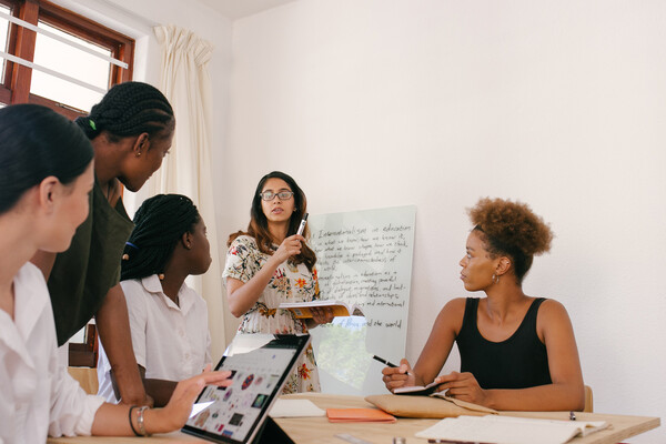 One person stands before a table with four other people, they are writing on a whiteboard while the other four people take notes and hold tablets.