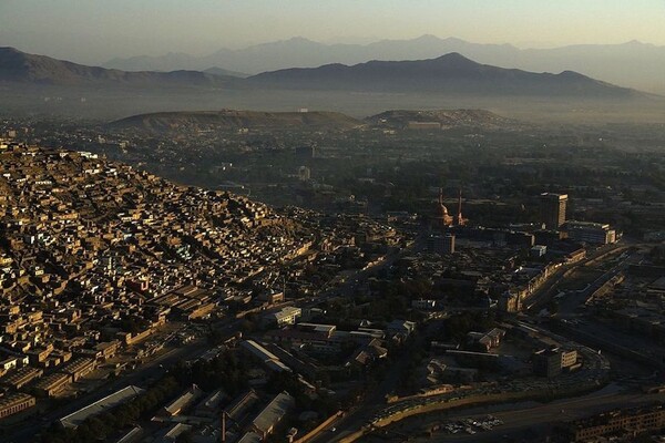 A city in the desert is seen from above, with brown structures dotting the valley and mountain ranges in the distance, some covered in haze.