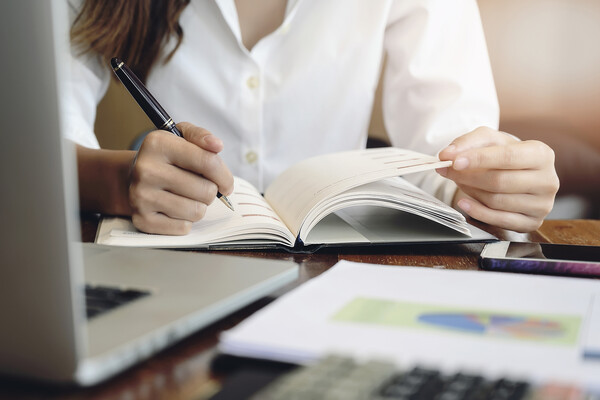 Person making marginalia in a journal in front of a laptop with graphs on paper on the desk.
