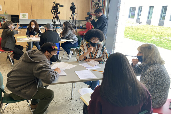 Masked adults sit four to a table with papers and folders during a brainstorming session.
