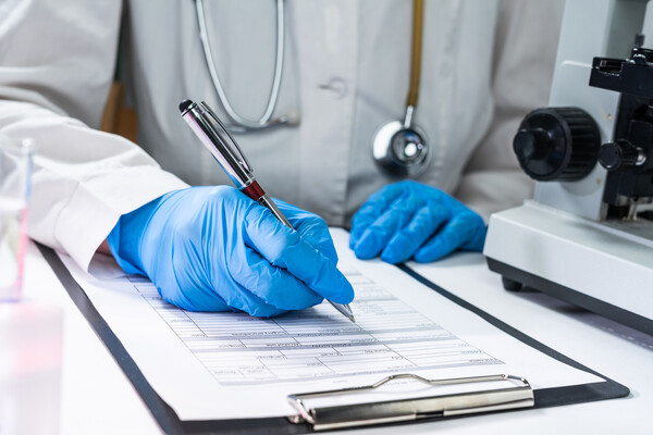 Doctor is white coat wearing stethoscope and latex gloves seated at desk filling out information on a clipboard.