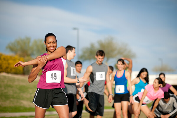 An African-American woman stretches her arms in the foreground; men and women stretch in the background. All are wearing running clothes with numbers on the front of their shirts.