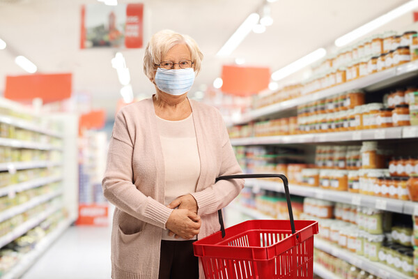 Senior citizen wearing a mask holding a shopping basket in a grocery store aisle.