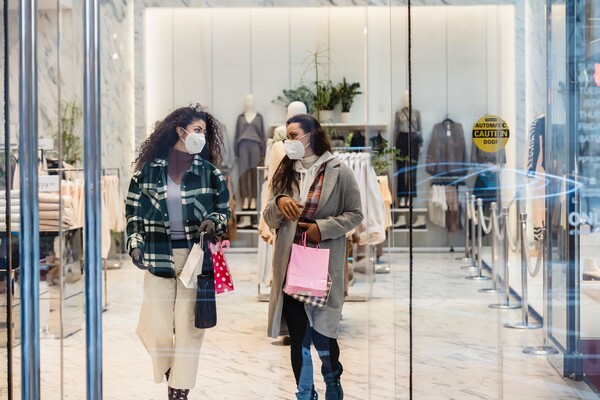 Two masked women holding shopping bags talk as they walk out of a store