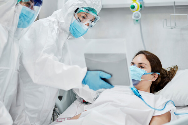 COVID-19 patient in a hospital bed wearing a mask with two medical personnel beside them in full PPE.
