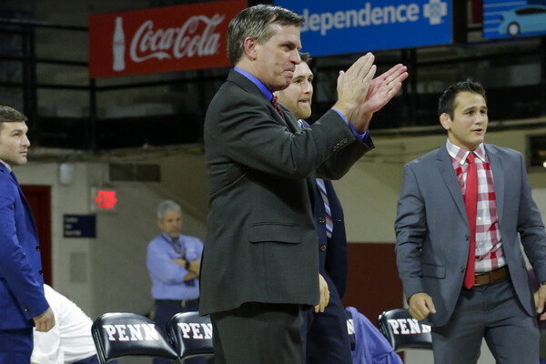 Roger Reina, wearing a suit and tie, claps his hands while watching a wresting match from the sidelines.