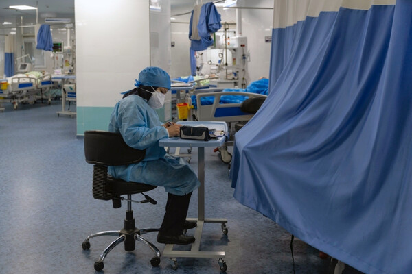 A nurse seated at a work station in a large hospital room with patient beds.