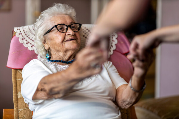 An elderly person, seated, holds hands with a caregiver standing over them.