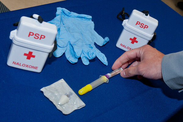 A hand holding a syringe on a table with latex gloves and two naloxone kits.