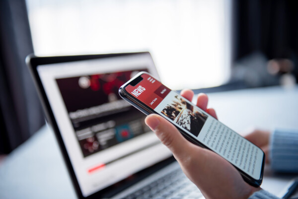 Person working on a laptop looking at their smartphone with a news feed on it.