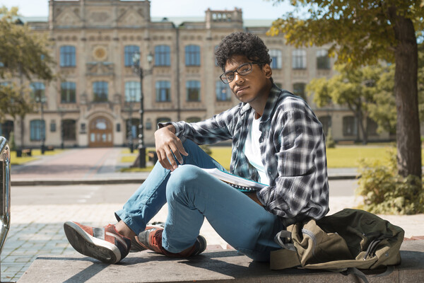 African American person sitting outside on a college campus with books and a backpack.