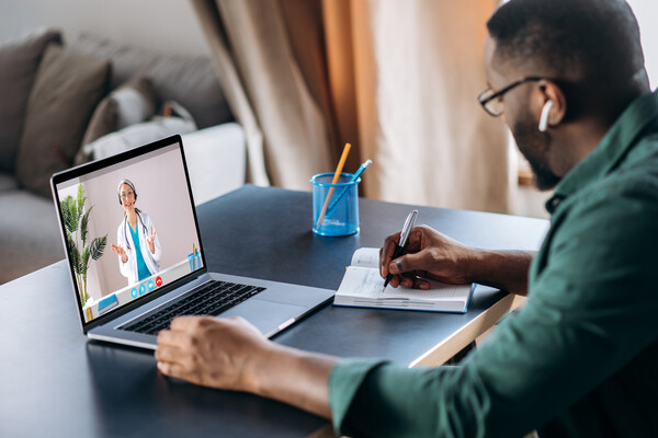 Person sitting at a laptop on a table taking notes having a telehealth appointment.