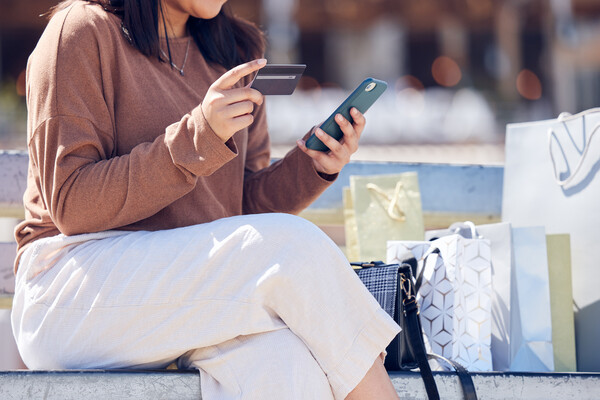 Person sitting on bench with a smartphone and credit card surrounded by shopping bags.