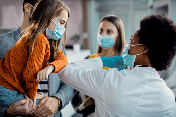 A family in a doctor’s office, one parent holds a young child who is elbow-bumping a masked doctor.