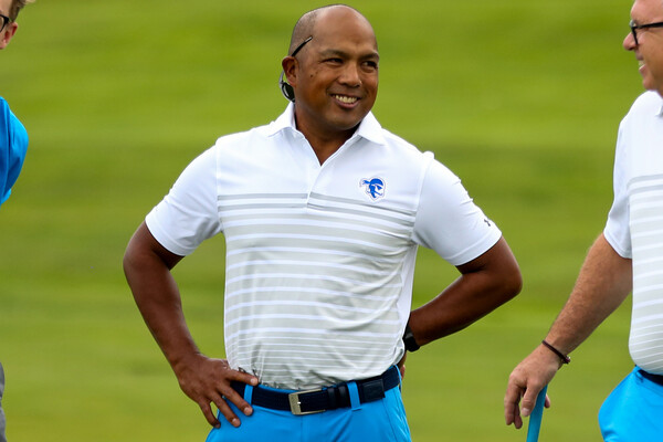Clay White, wearing a Seton Hall shirt, stands on a golf course whith his hands on his hip, while standing in between two people.