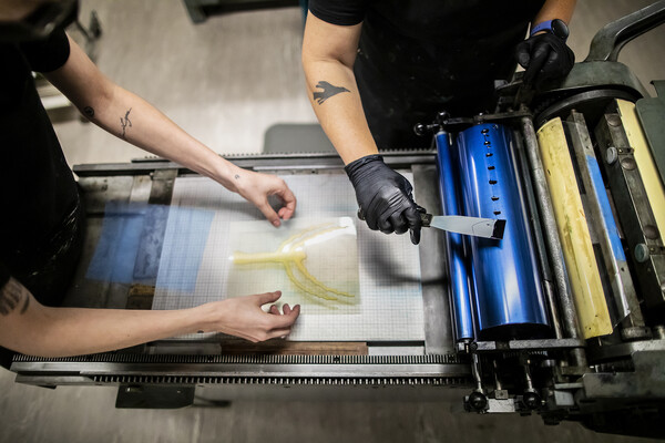 two sets of arms over a hand-operated printing press, one set with gloved hands putting ink on a metal cylinder and the other placing a printing plate with an image of a tree without leaves on the flat surface in front of the cylinder