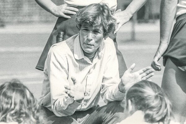 Anne Sage kneels down to address her players, who are seated on the field. Two players stand behind her.