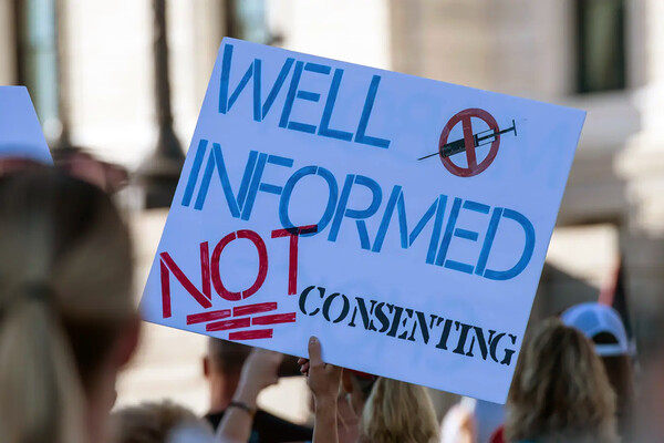 A person holds a sign during a protest that reads WELL INFORMED NOT CONSENTING.