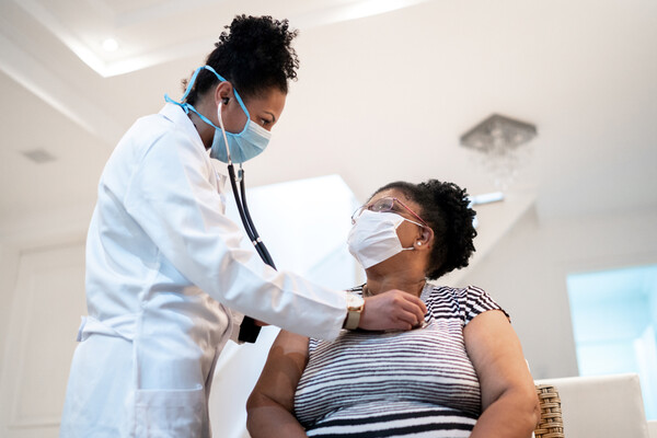 A masked nurse checks the heartbeat of a seated masked patient.