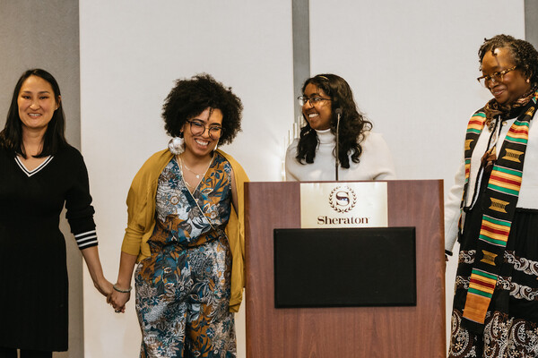 Zhanar Beketova, Krista L. Cortes, Nesha Subramaniam, and Sharon Smith hold hands at the podium