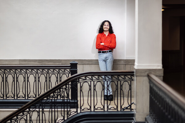 A student in a red long sleeved shirt stands with arms crossed leaning against the wall atop a staircase.