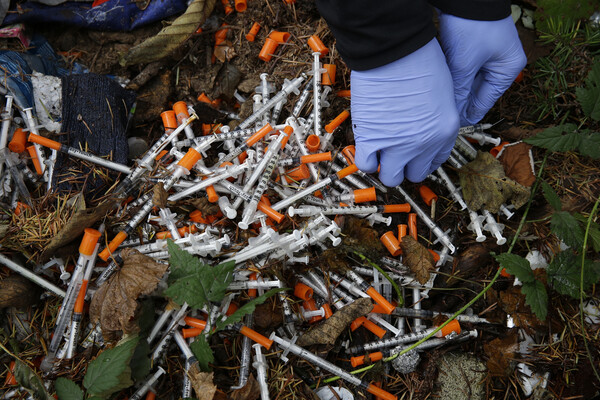 Gloved hands clean up a collection of needles at an encampment for unhoused people.