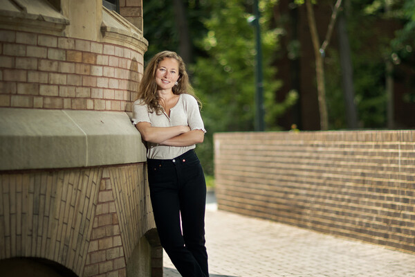 History Ph.D. candidate Arielle Alterwaite leans up against a light brown brick building with her arms crossed, smiling at the camera.