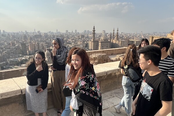 A group of students take pictures on a rooftop deck. The city of Cairo, with its skyscrapers and minarets, are visible in the background