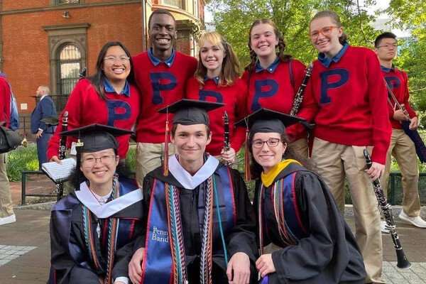 Five people wearing red sweaters with an applique "P" holding clarinets. In front of them, three graduating bandmates sport gowns and regalia