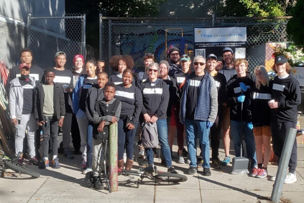 A group of people standing on the sidewalk in front of a fence with a Clean, Green & Serene sign.