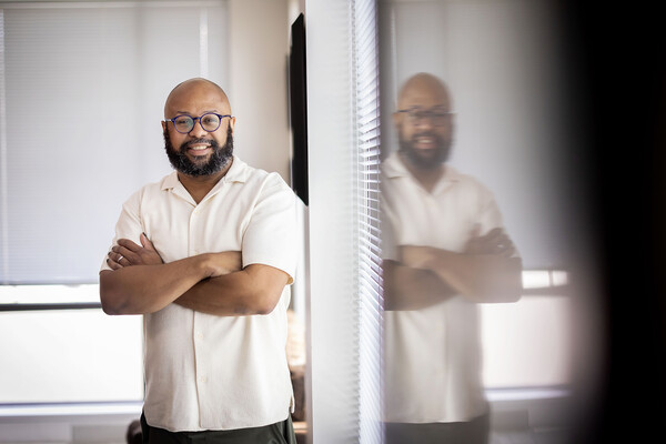 Desmond Patton stands with arms crossed in front of a window, which mirrors his image