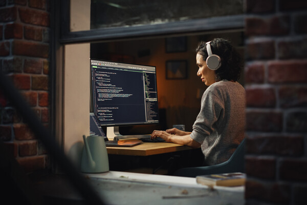 A person working at home on their laptop with headphones as seen through an open window.