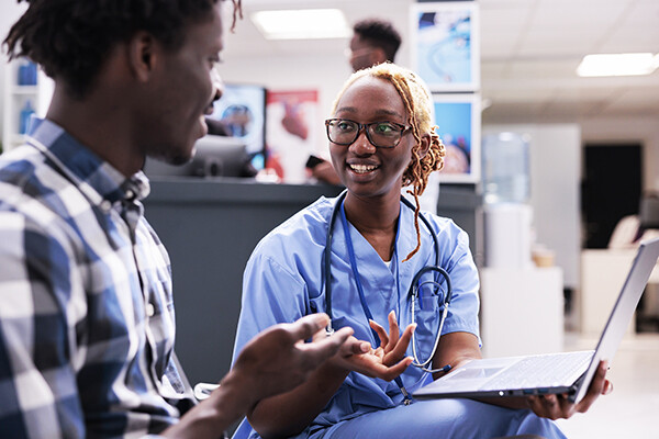 A doctor, at right holding a laptop, speaks witn a patient about his health.