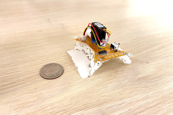 A centimeter-scale quadruped robot sitting on a desk next to a a quarter for scale