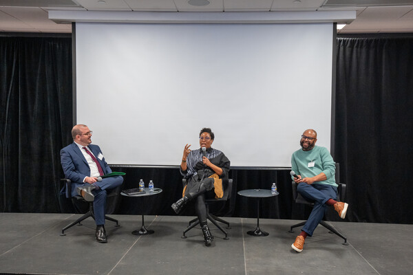 A stage with three chairs, two tables, and a blank screen; Bauermeister and Patton look on as Cogburn speaks. 