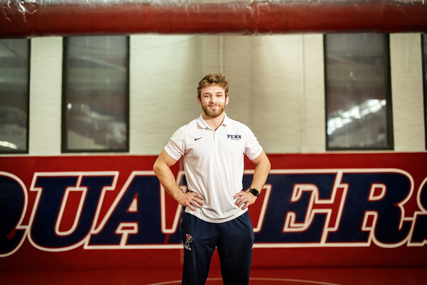 Wrestling Adam Thomson stands in the wrestling room at Penn, hands on hips, with a sign reading Quakers behind him.