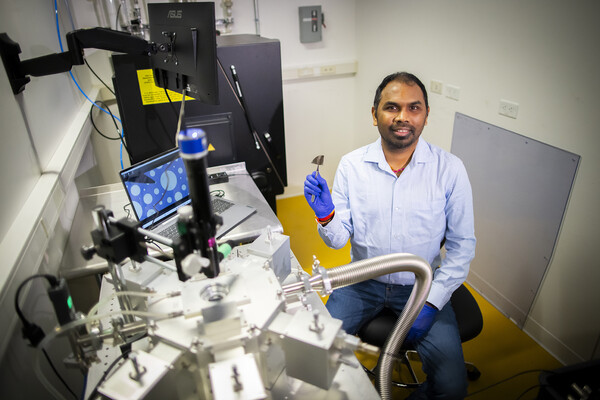 Researcher sits in a laboratory filled with equipment, holding up a small, metallic-looking sample for examination.