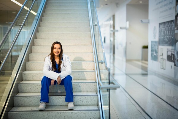 Katelyn Candido sitting on a staircase, smiling and facing forward, wearing a white coat and blue scrubs in the Perelman School of Medicine