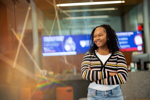 Mikhaidia Miller wearing a striped sweater and standing with arms crossed, smiling and facing sideways, in the Penn First Plus office.