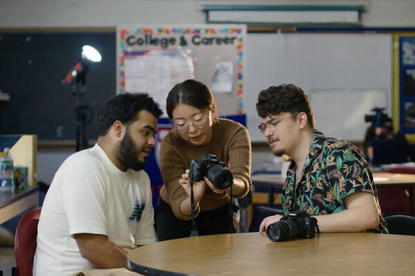 The two members of Lensbright looking at a camera with a student in a classroom.