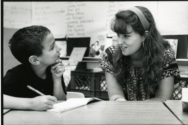 An elementary school student (left) and Penn affiliate (right) are interacting while the student writes in his booklet, in a classroom setting.