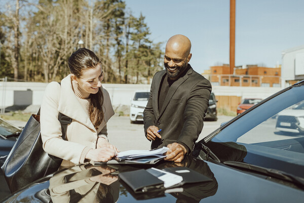 A person signing paperwork from a salesman at an outdoor car lot.