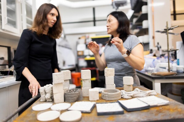 A tabletop display of off-white tiles in the foreground. Dorit Aviv and Shu Yang are standing behind it.  