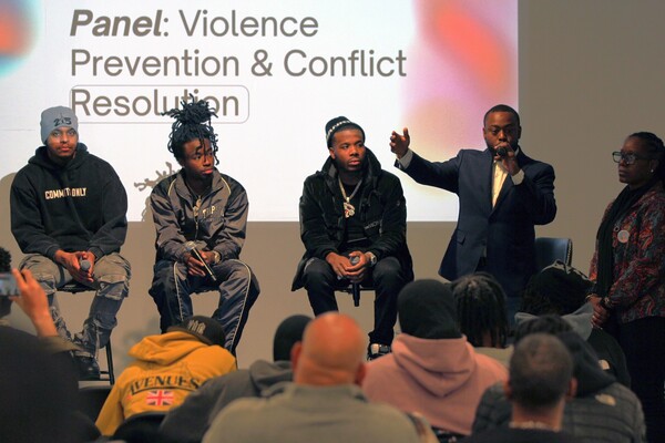 (from left to right): Social media creators Manny 215 [Manny Clark], Prince Dre, Fis Banga, and Ant Brown on stage with facilitator Tamara Russell at the Creators for Community Safety Summit. Brown is speaking and holding a microphone. The slideshow in the background reads: "Panel: Violence Prevention and Conflict Resolution"