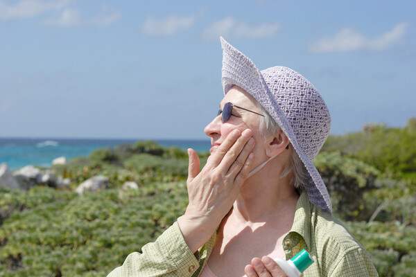 An older person in a hat outdoors applying sunscreen.
