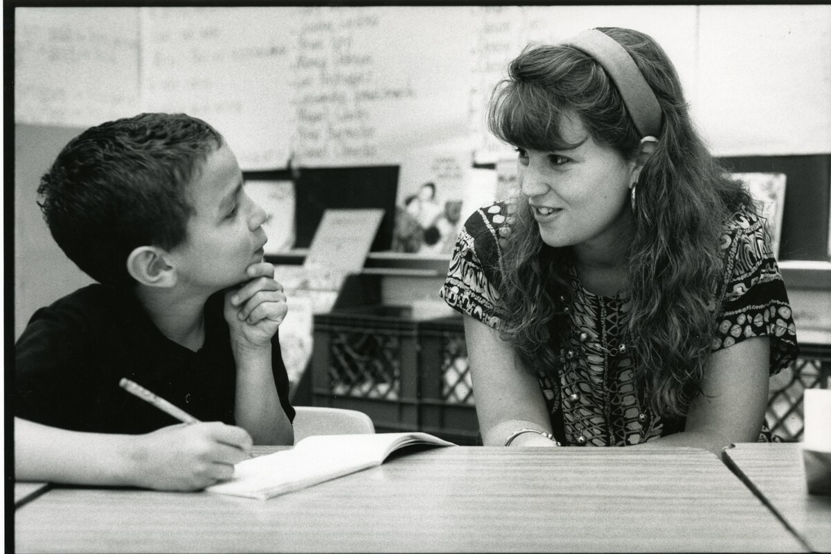An elementary school student (left) and Penn affiliate (right) are interacting while the student writes in his booklet, in a classroom setting.