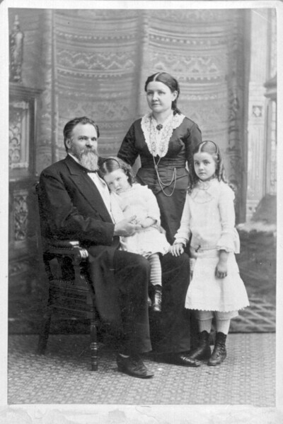 father sitting in chair with daughter on his lap, mother and other daughter standing next to the chair