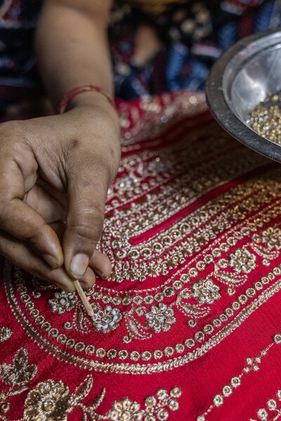 The hand of an Indian woman worker carefully applies small jewels to a piece of red cloth in an intricate design.